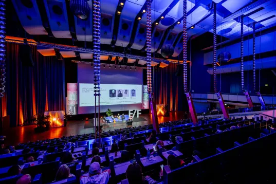 Modern auditorium with stage, lit in blue and orange, audience watching.