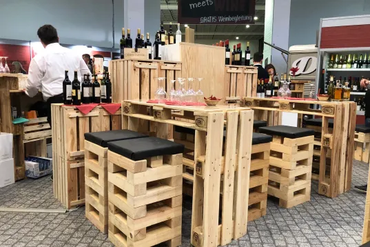 Wooden pallet bar setup with wine bottles, stools, and glasses in a market setting.
