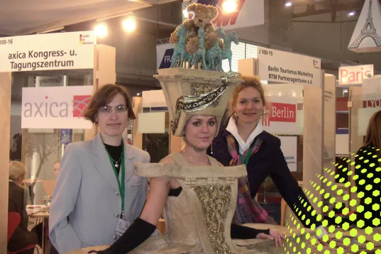 Drei Frauen posieren auf einer Messe, eine trägt ein auffälliges Kostüm mit einer Bärenfigur.