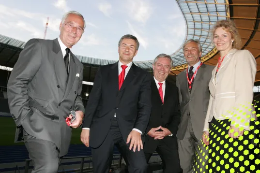 Fünf Personen im Anzug stehen lächelnd in einem Stadion unter freiem Himmel.