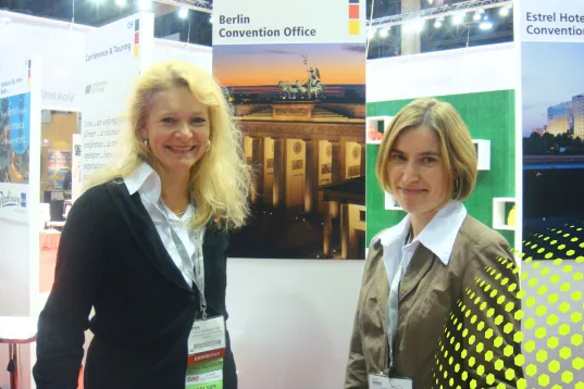 Zwei Frauen stehen lächelnd auf einer Messe vor Messeständen mit Berlin-Postern.