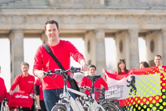Lächelnder Mann mit Fahrrad vor dem Brandenburger Tor, umgeben von Menschen in Rot.