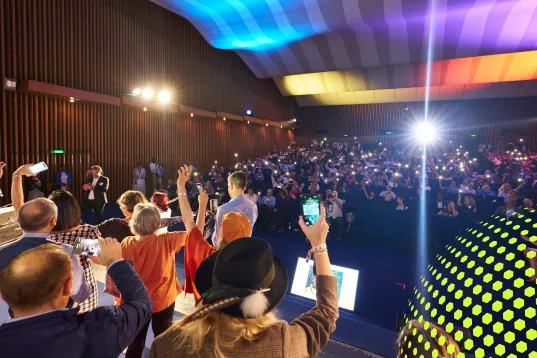 Menschen auf einer Bühne mit Publikum in einem bunt beleuchteten Saal