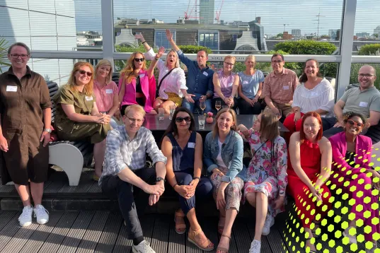 Gruppe von Menschen sitzt und steht lachend auf einer sonnigen Dachterrasse.