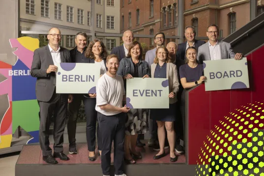 Gruppe von Menschen hält Schilder mit „Berlin Event Board“ in modernem Innenraum.