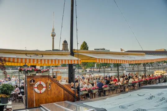 Outdoor riverside café with people dining under striped awnings, city skyline in background.