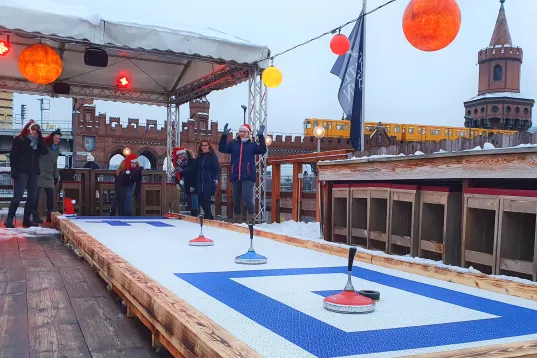 People are playing curling on an outdoor rink, with colorful lanterns hanging above it.
