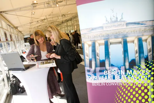 Zwei Frauen sprechen an einem Messestand mit Berlin-Werbeplakat im Hintergrund.