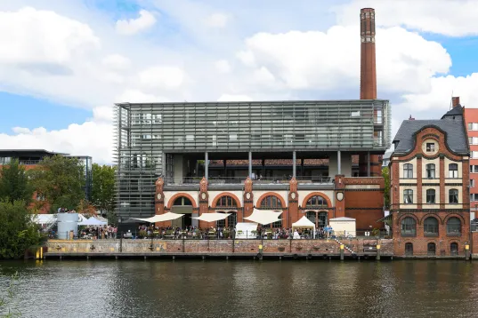 A brick building with a glass façade and a chimney by the river, with people and tents in front of it.