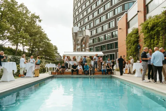 People socializing around a rooftop pool beside a modern building on a cloudy day.