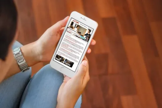 Person holding a smartphone while reading news on a wooden floor.