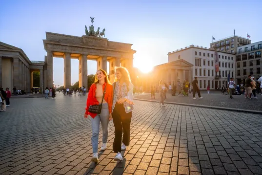Zwei Frauen spazieren vor dem Brandenburger Tor bei Sonnenuntergang.