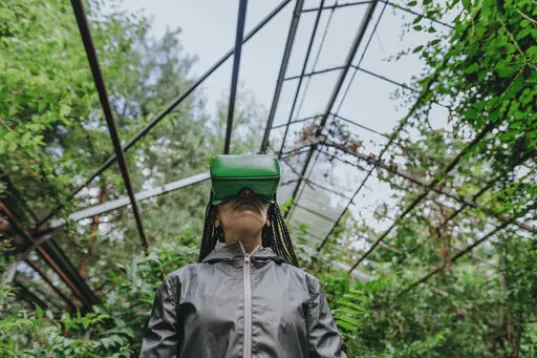 A woman with braided hair wears VR glasses in a greenhouse.