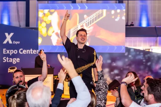 A saxophonist in a jubilant group, with the words ‘X Expo Expo Stage’ visible in the background.
