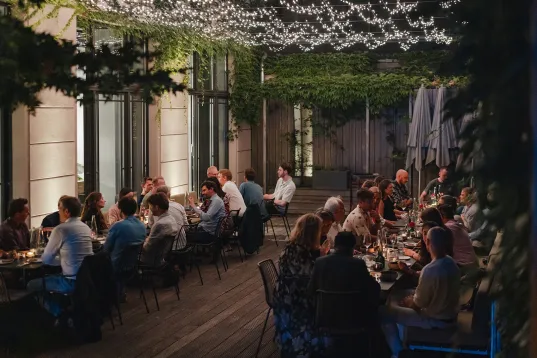Menschen essen abends auf einer Terrasse unter Lichterketten und grünen Pflanzen.