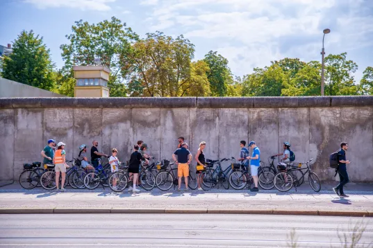 Group of people with bicycles gathered beside a concrete wall on a sunny day.