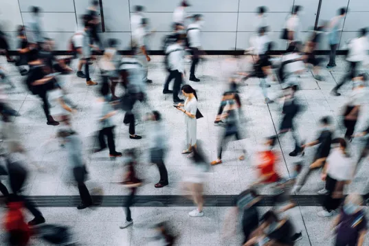 Young Asian woman using a smartphone, surrounded by commuters rushing past in a underground station during rush hour.