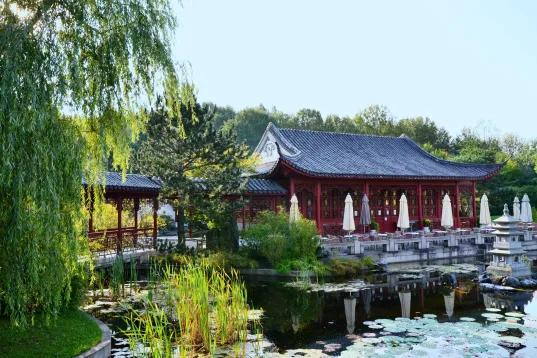 The photo shows a red Asian pavilion with sun umbrellas and green surroundings under a clear sky.