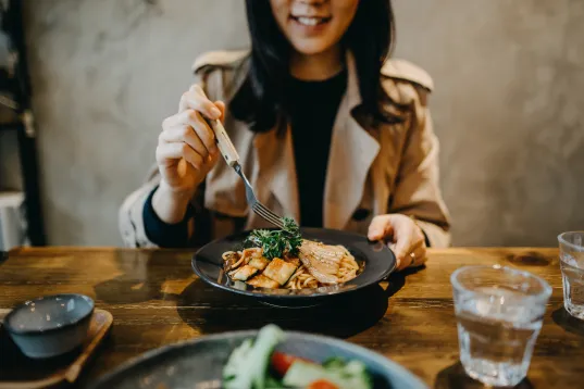 Frau lächelt beim Essen in einem gemütlichen Restaurant, Teller mit Speise vor ihr.