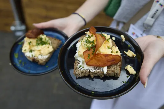 Person holding two plates with bread, scrambled eggs, cream cheese, chives and baked vegetable crisps.