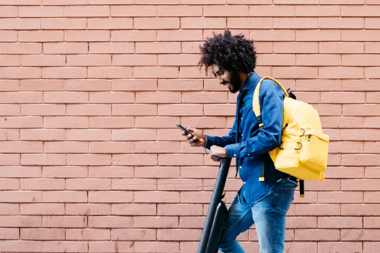 Man with yellow backpack on scooter looking at phone by a brick wall.