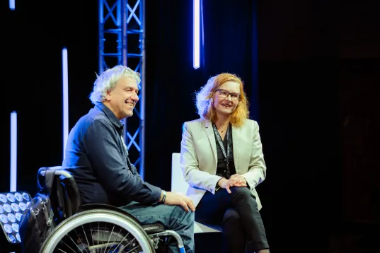 On stage, a man in a wheelchair and a woman are seated. Both are smiling and looking in the same direction