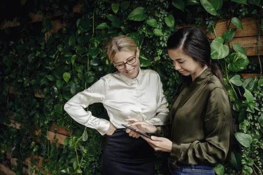 Two women in business attire stand in front of an ivy-covered wall and look at an Tablet with smiles on their faces.