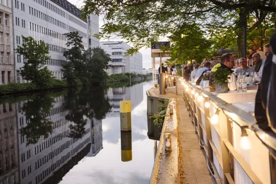 Blick auf den Kanal am Estrel Berlin mit beleuchtetem Uferweg, spiegelnden Gebäuden und Bäumen im Wasser bei Abendlicht