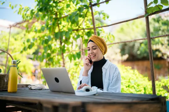 Woman sitting outdoors at a table with a laptop, smiling and talking on the phone