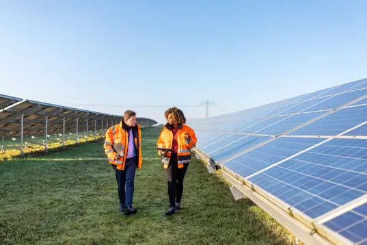 Male and female engineers working at solar power plant. Two technicians in reflective clothing walking between rows of photo…