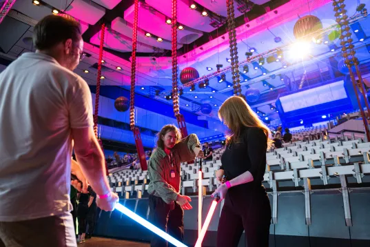 People playing with glowing lightsabers in a colorful, empty auditorium.