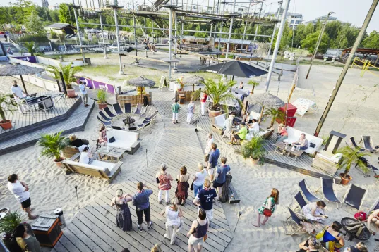 People socializing at a sunny outdoor beach bar with loungers and palm trees.