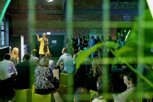 A speaker addresses an audience in a modern, indoor event space with green accents.