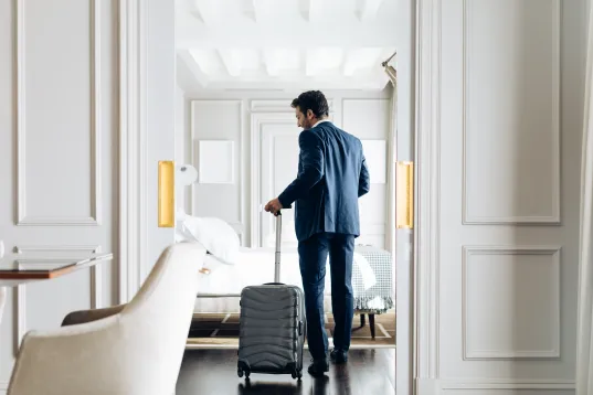 Mann im Anzug betritt mit Rollkoffer ein elegantes Hotelzimmer.