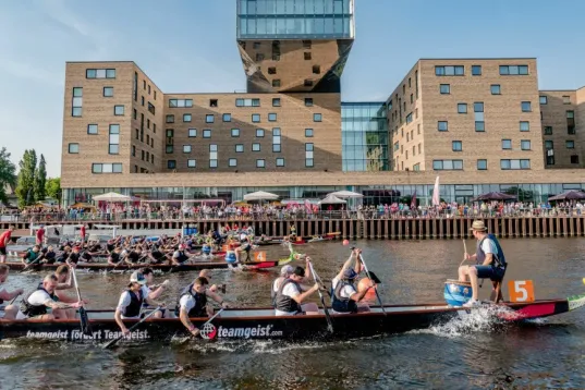 Two dragon boat teams racing on a river in front of a modern brick building.