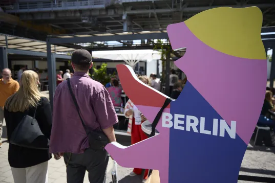 Buntes Bären-Logo mit „Berlin“; Menschen gehen im sonnigen Außenbereich vorbei.