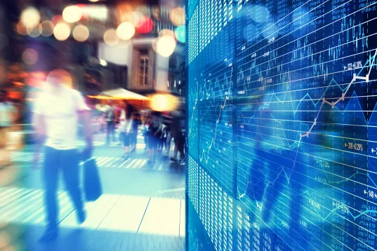 Menschen laufen an einer digitalen Anzeigetafel mit Finanzdaten und Diagrammen vorbei.