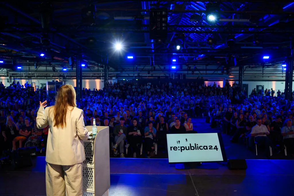 Rednerin spricht auf einer Bühne vor großem Publikum bei einer Konferenz.